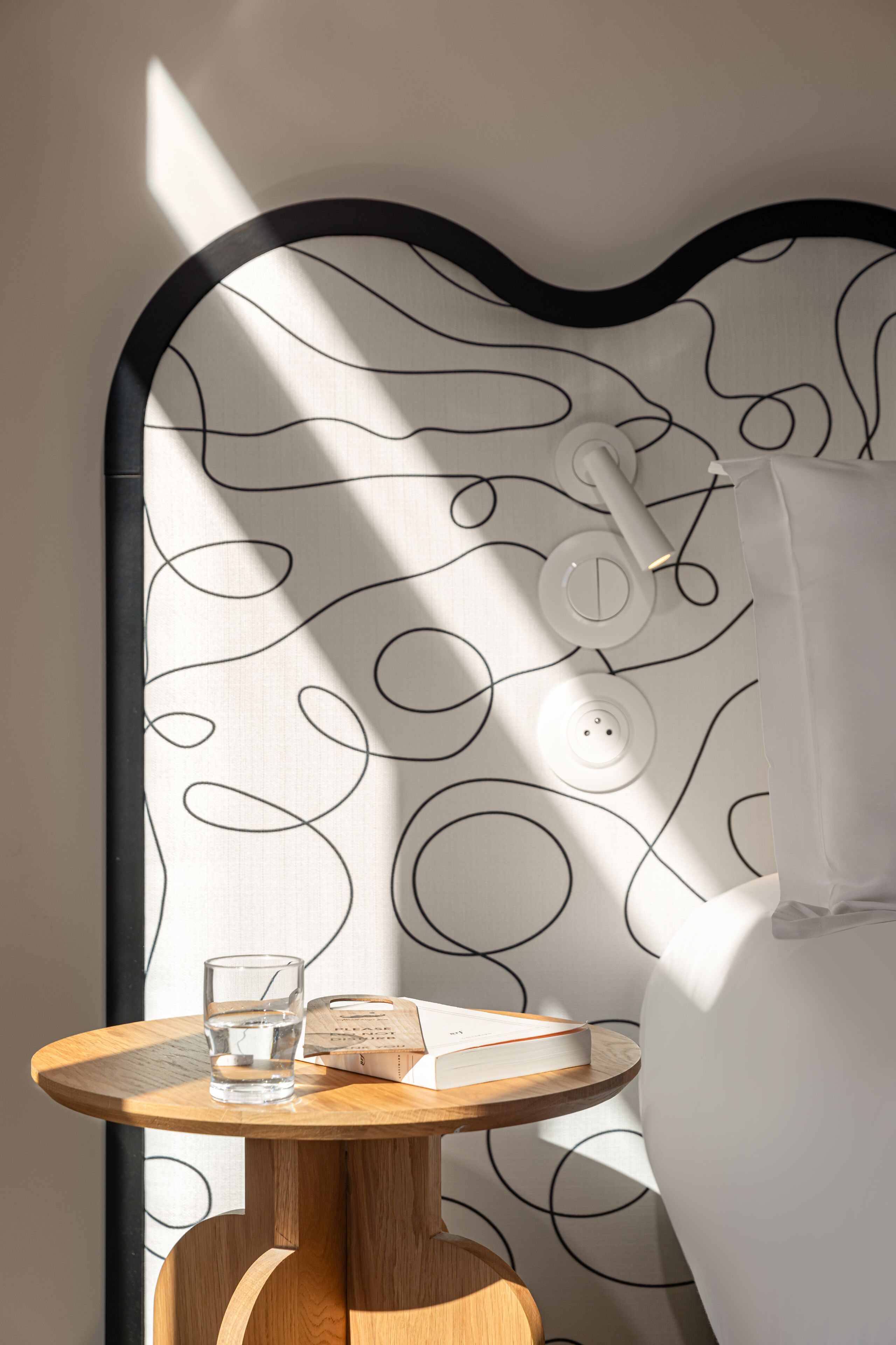 A bathroom with a modern design, featuring a round wooden table with a glass of water and a book, a white shower curtain with a black abstract pattern, and a wall-mounted towel rack.