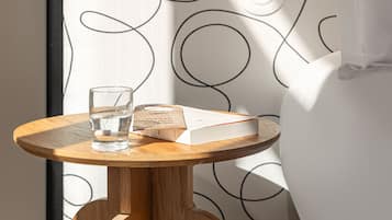 Un baño con diseño moderno, que incluye una mesa redonda de madera con un vaso de agua y un libro, una cortina de ducha blanca con un motivo abstracto negro, y un perchero de toallas montado en la pared.