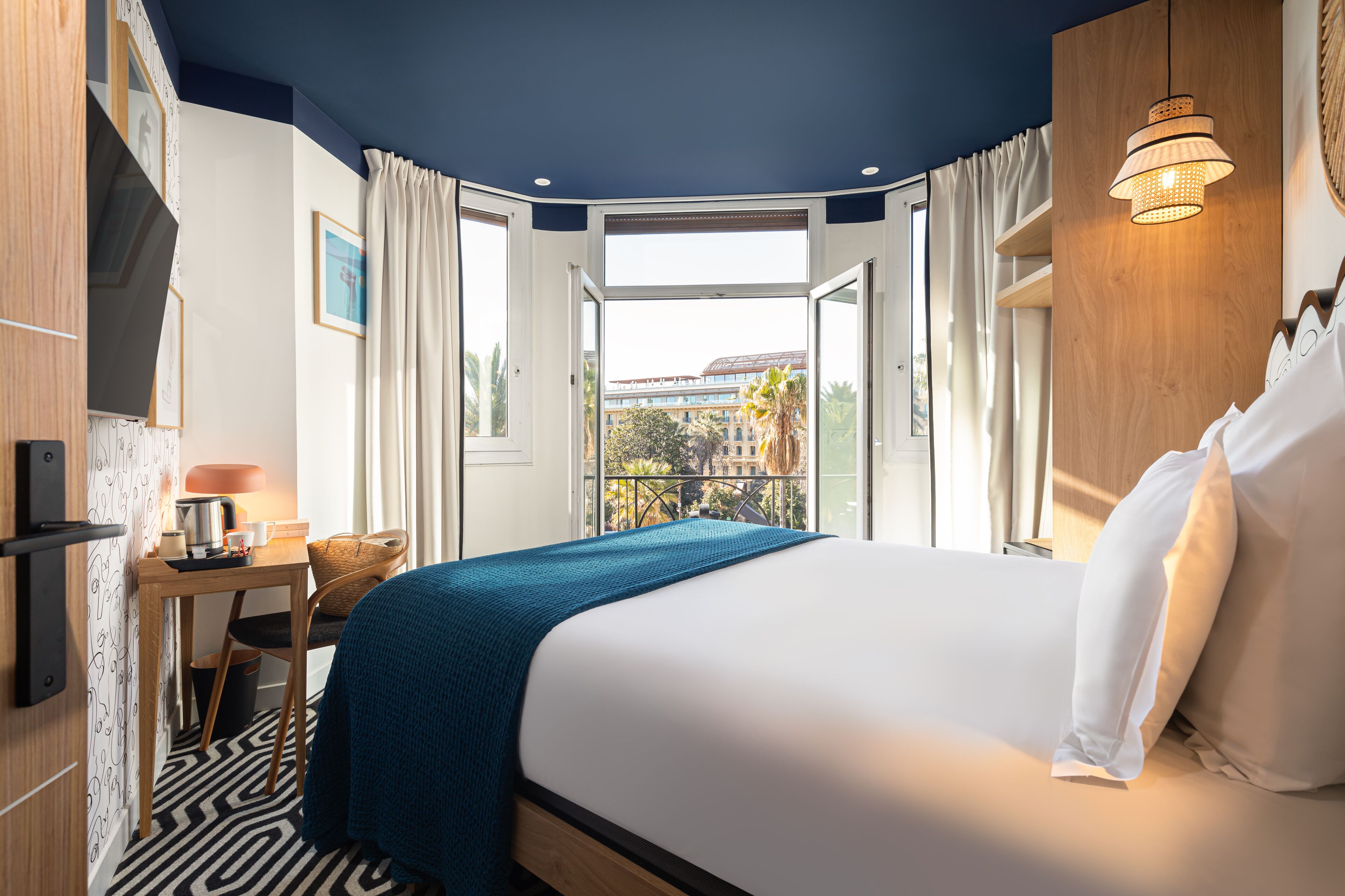 A hotel room with a large bed, a desk with a lamp, a chair, and a view of the city through an open door.
