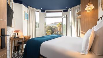 A hotel room with a large bed, a desk with a lamp, a chair, and a view of the city through an open door.