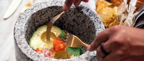 A person using a mortar and pestle to prepare a dish with avocado, tomatoes, and cilantro.