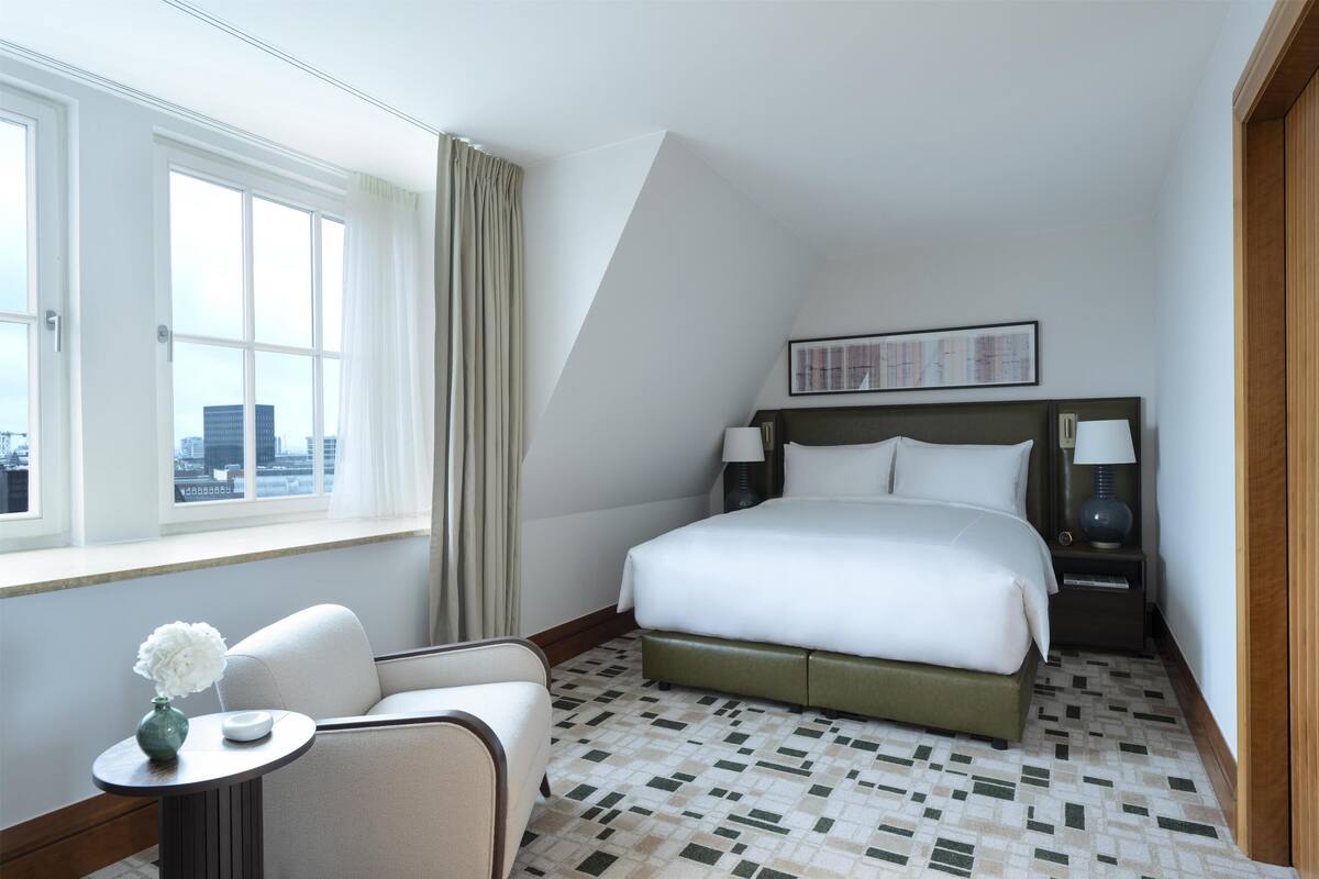 A hotel room with a bed, a chair, a nightstand, a lamp, and a window with curtains.
