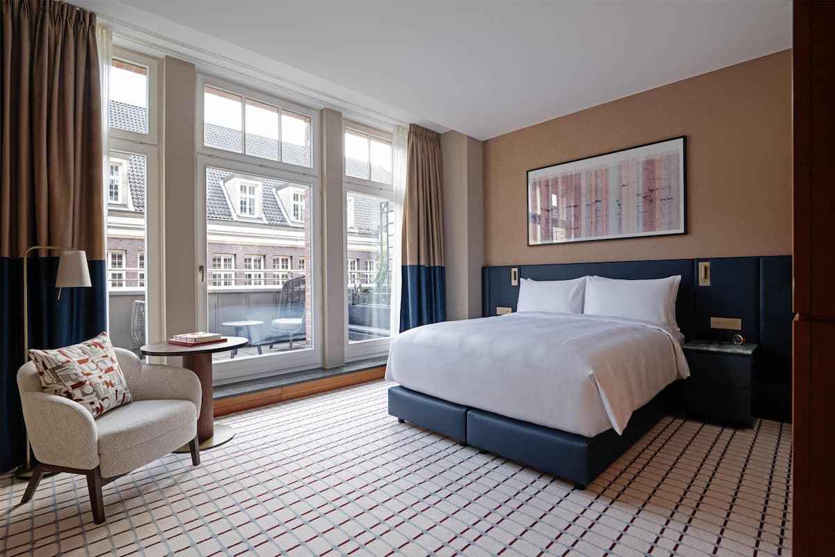 A hotel room with a large bed, a chair, a small table, and a window with curtains.