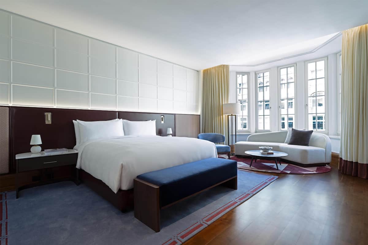 A hotel room with a large bed, a sofa, a bench, a desk with a lamp, and a view of the city through large windows.