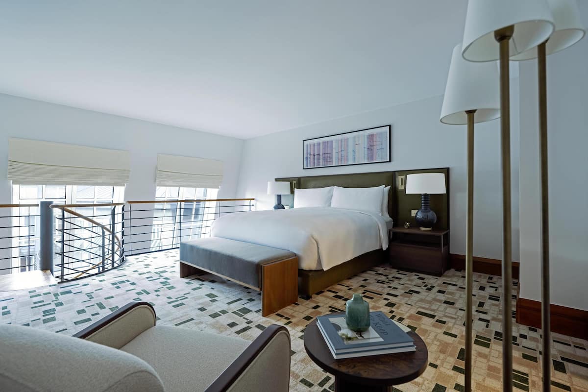A hotel room with a bed, a nightstand, a lamp, a chair, a small table, and a view of the outside.