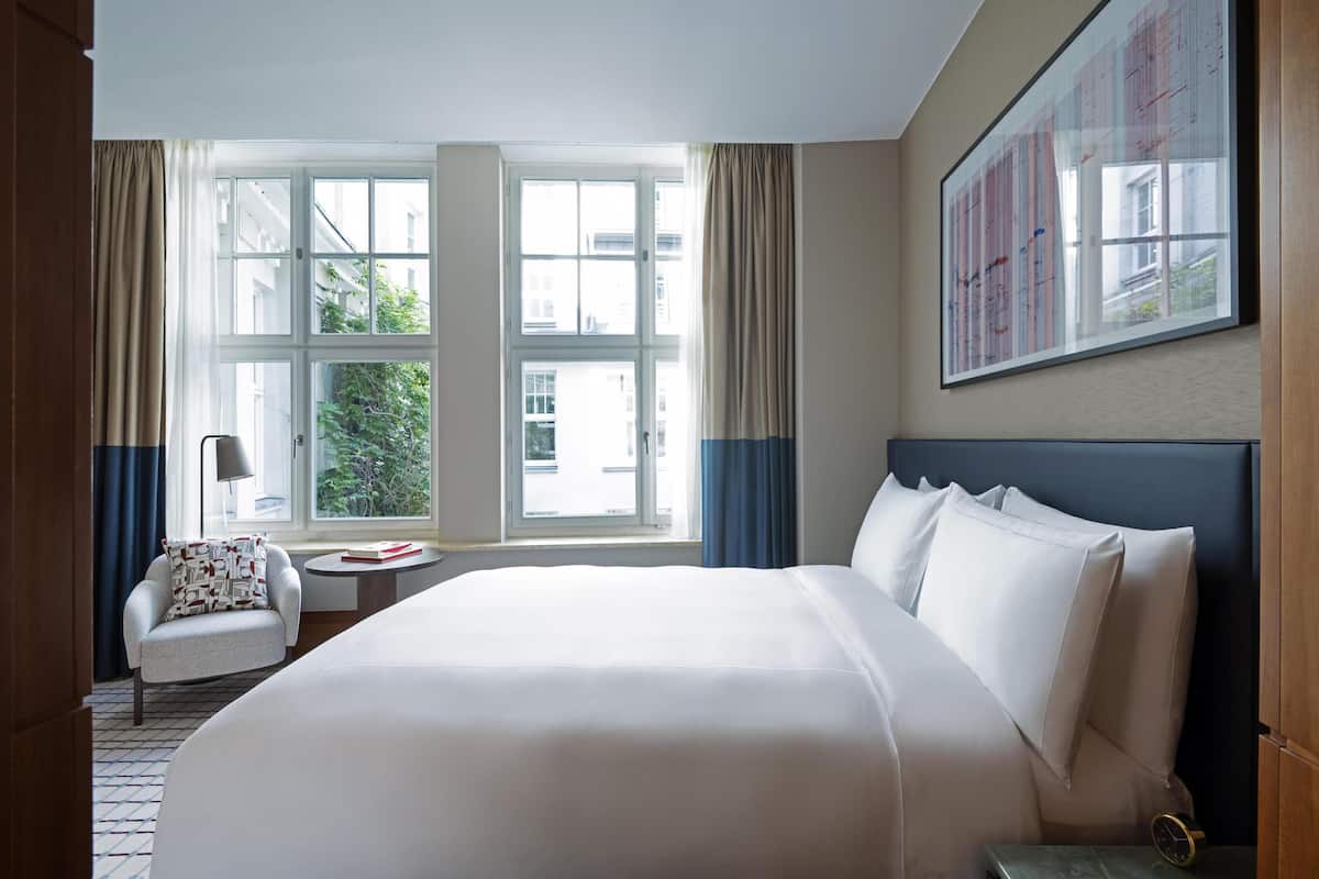A hotel room with a large bed, a chair, a small table, and a window with curtains.
