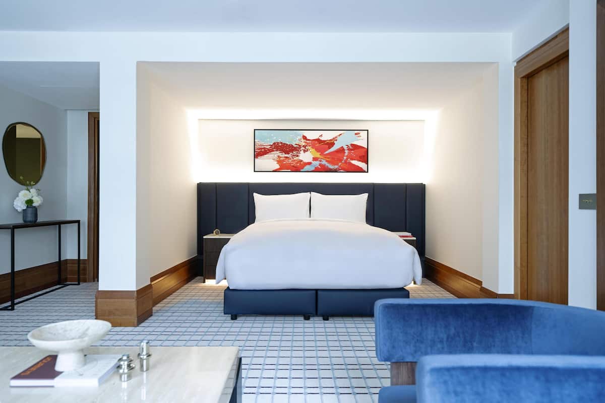 A modern hotel room with a large bed, a bedside table, a chair, and a wall-mounted artwork.
