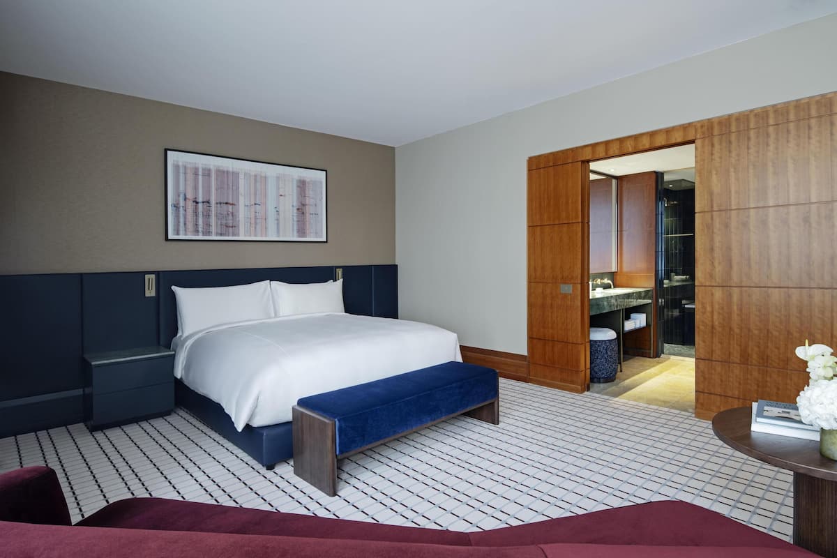 A hotel room with a bed, a bench, a small table with a vase, a mirror, and a bathroom visible through an open door.