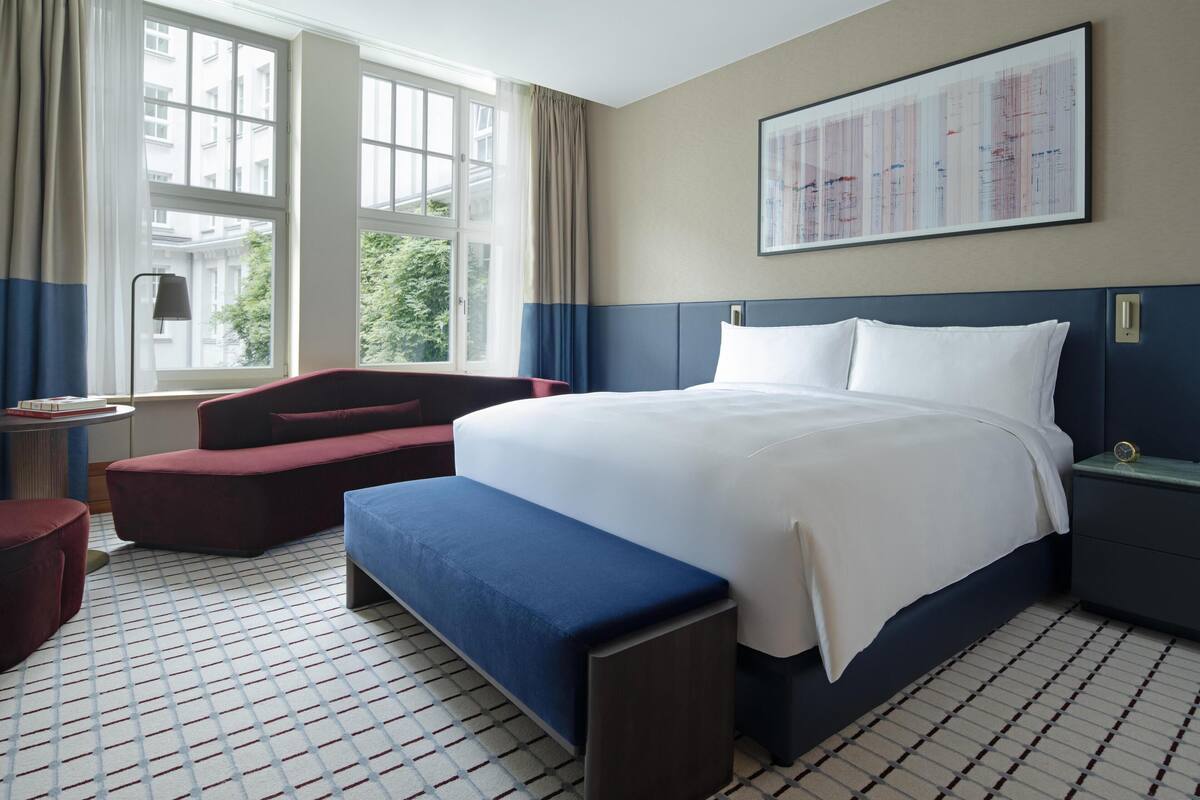 A hotel room with a large bed, a bench, a desk, and a window with curtains.
