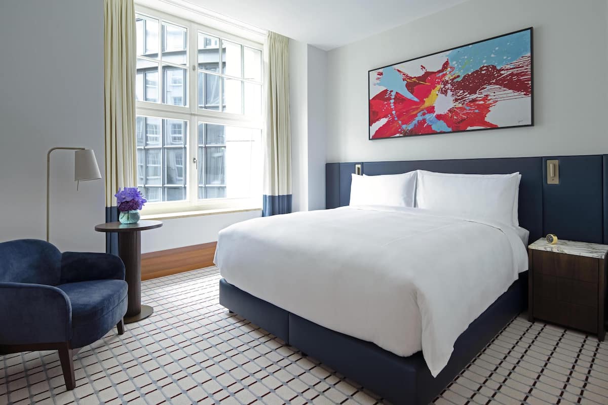 A hotel room with a large bed, a side table with a lamp, a chair, and a window with curtains.