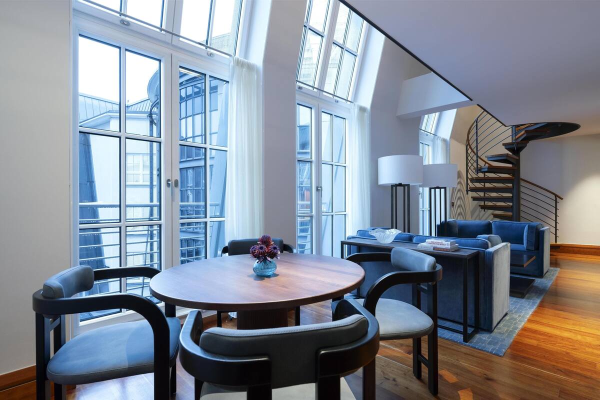 A modern living room with a round wooden table, blue upholstered chairs, a blue sofa, and a spiral staircase.