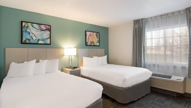 A hotel room with two beds, a nightstand with a lamp, a window with sheer curtains, and two abstract paintings on the wall. - MainStay Suites Raleigh - Cary (Cary)