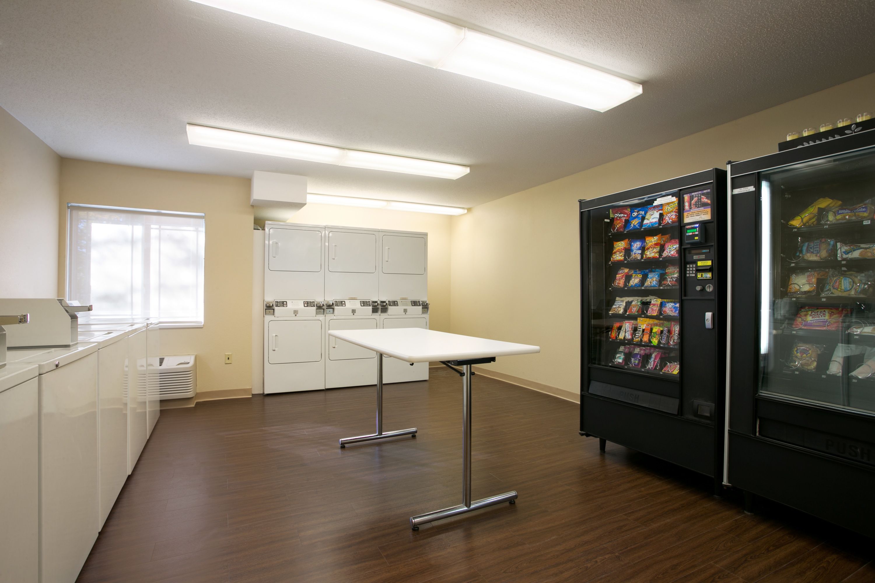 A kitchen with a white table, a refrigerator stocked with snacks, and a vending machine.