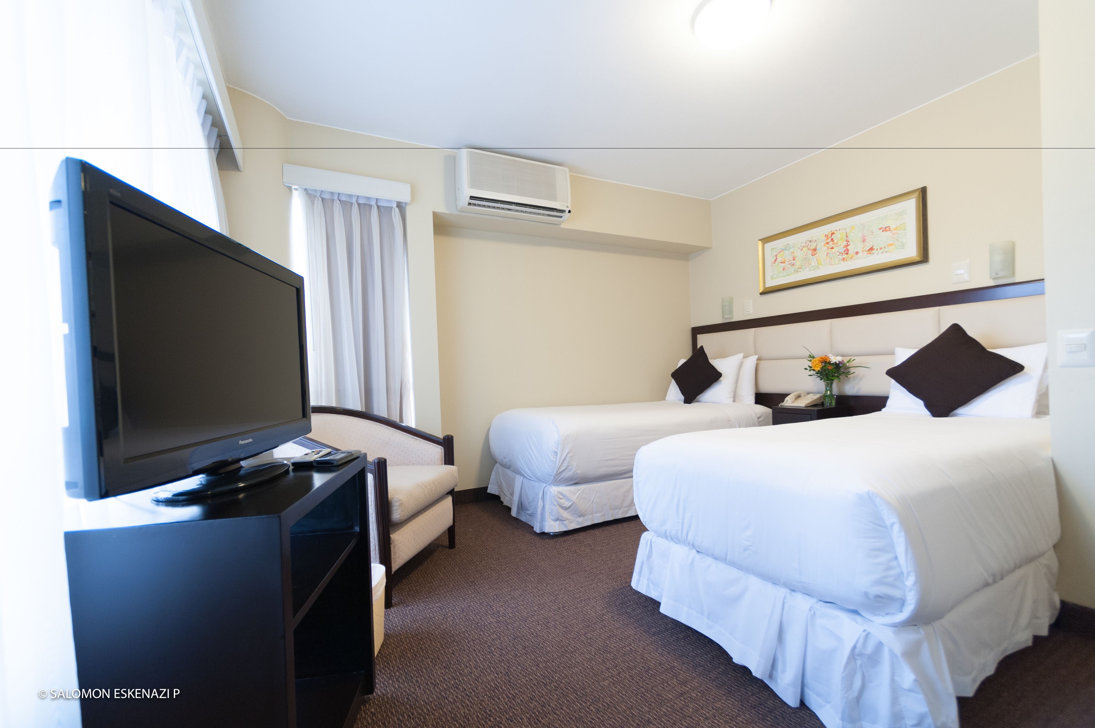 A hotel room with two beds, a TV, a desk, and a window with curtains.