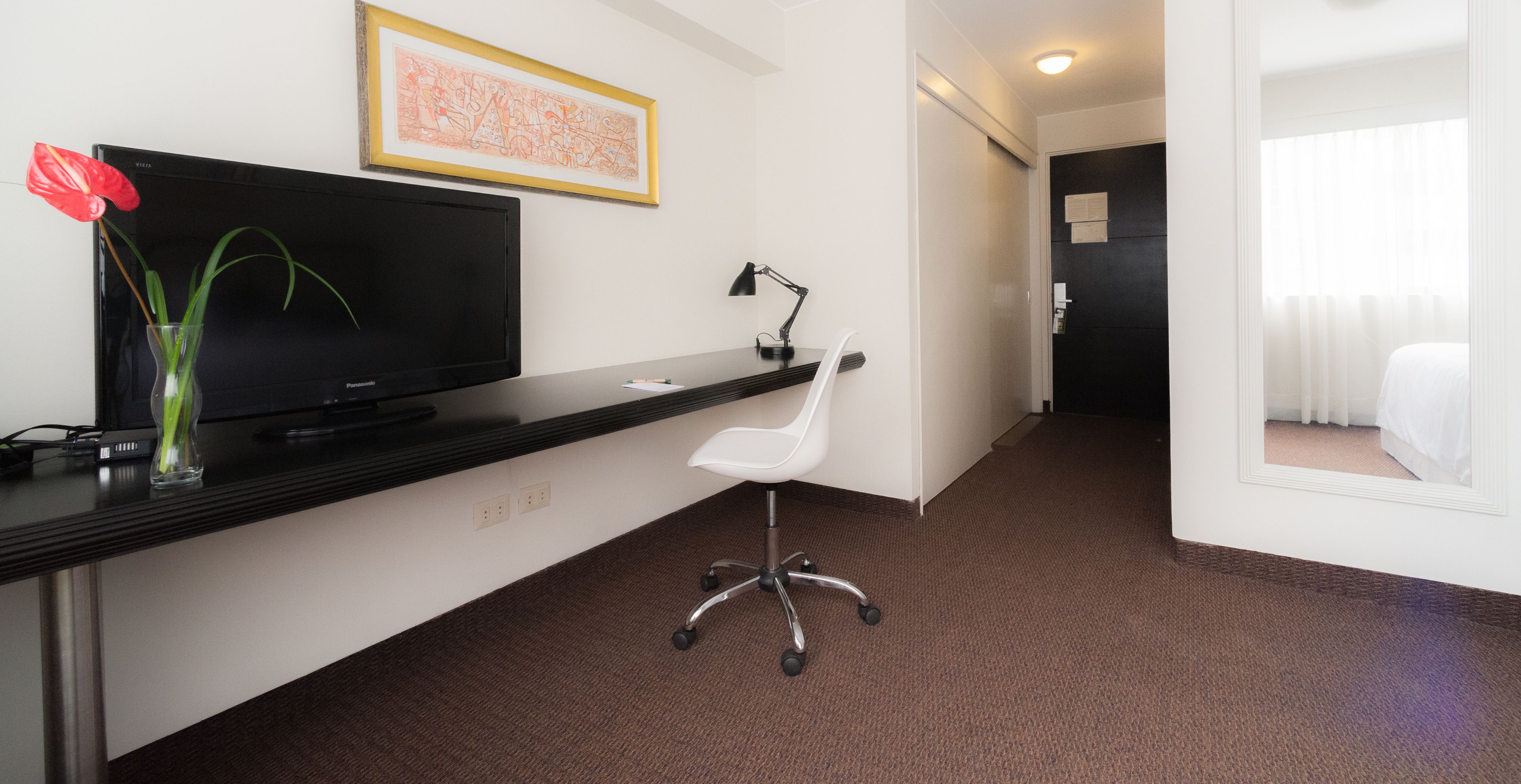 A hotel room with a flat-screen TV, a desk with a lamp, a white chair, and a bed.
