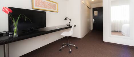 A hotel room with a flat-screen TV, a desk with a lamp, a white chair, and a bed.