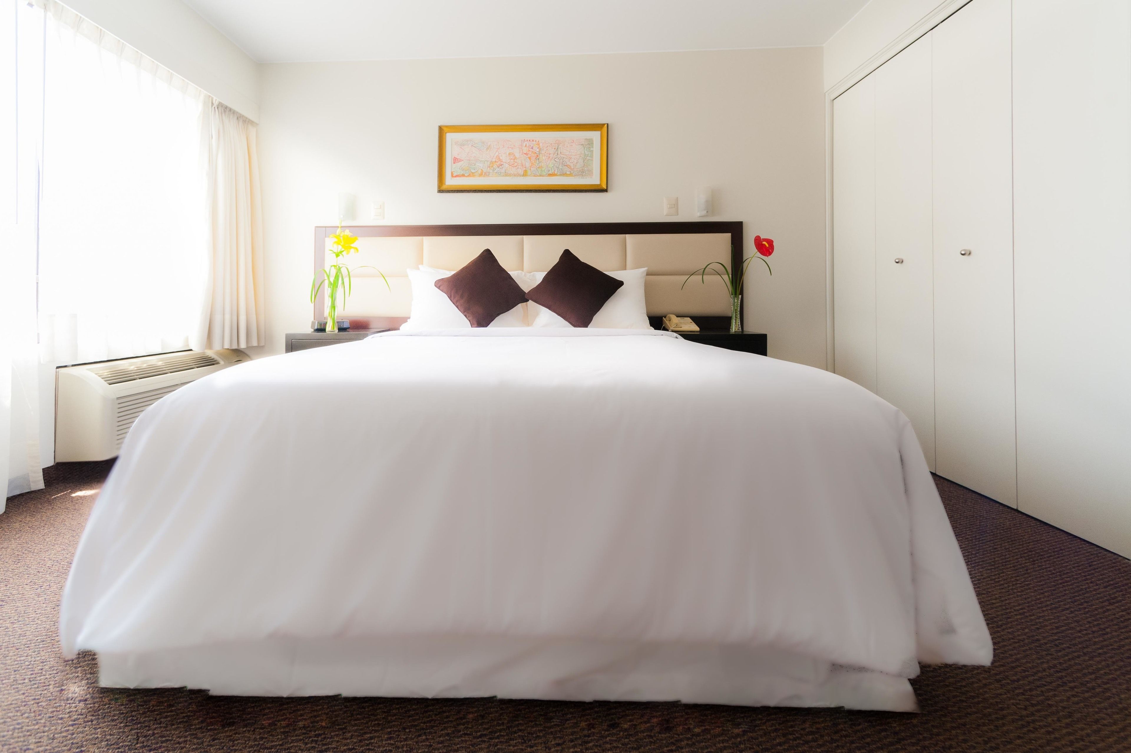 A hotel room with a large bed, a bedside table with a lamp, a framed picture on the wall, and a window with curtains.