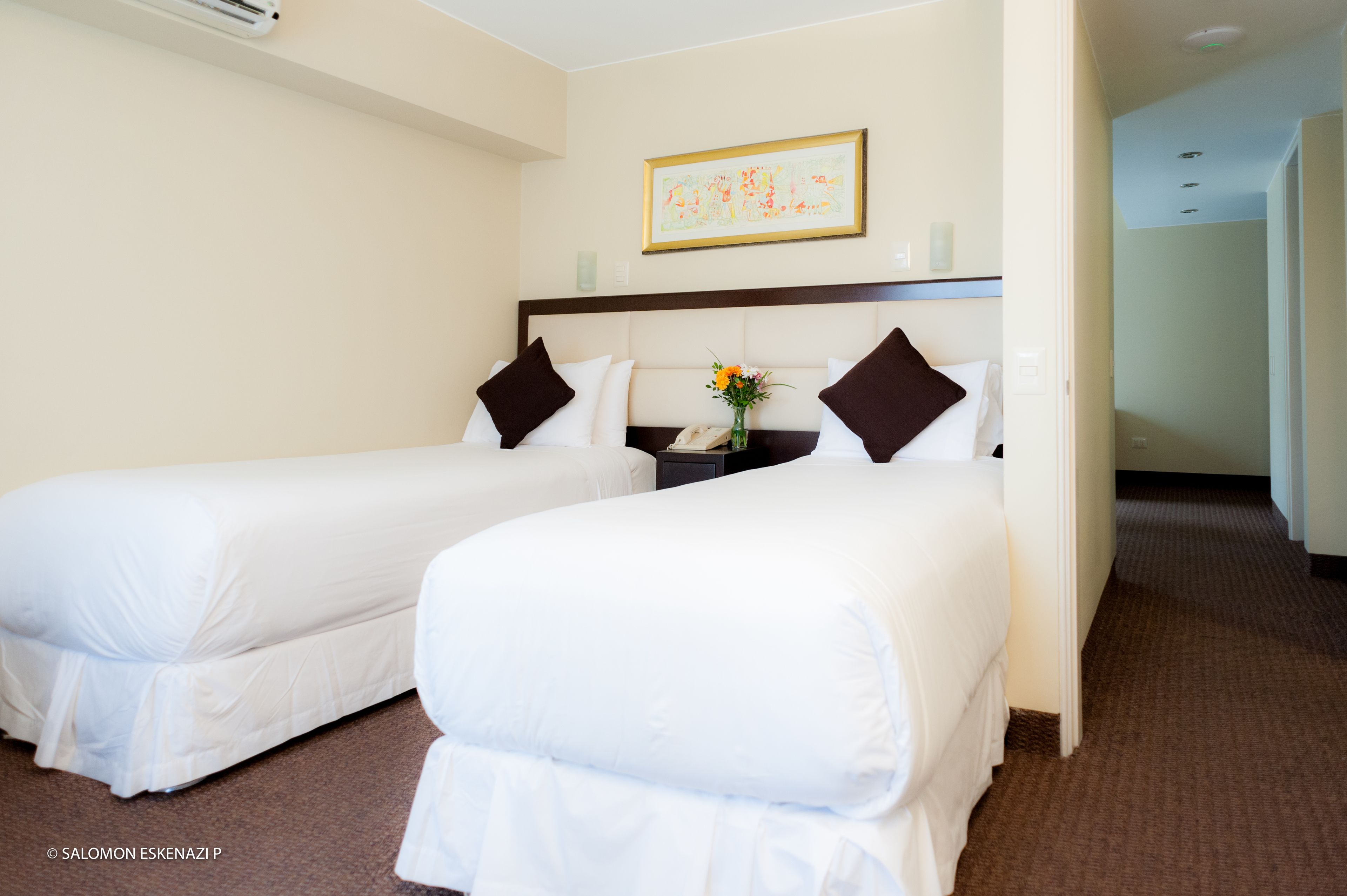A hotel room with two single beds, a nightstand with a vase of flowers, and a framed artwork on the wall.
