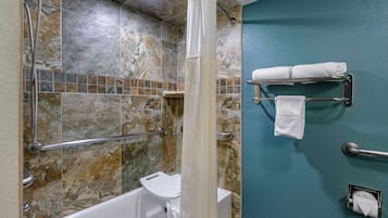Ein Badezimmer mit Badewanne, Dusche und Haltegriffen. Handtücher befinden sich an einem Wäscheständer, und es ist eine Toilettenpapierhalterung vorhanden.