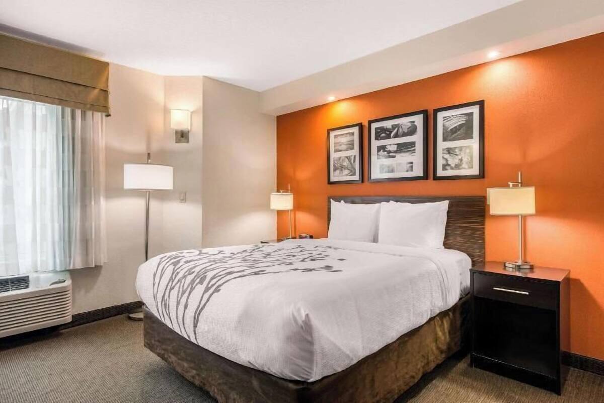 A hotel room with a bed, bedside lamps, a wall-mounted air conditioner, and framed pictures on the wall.