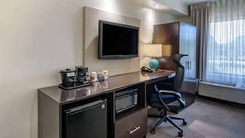 A hotel room with a flat-screen TV mounted on the wall, a desk with a coffee maker, microwave, and a lamp. There is a chair and a small refrigerator.