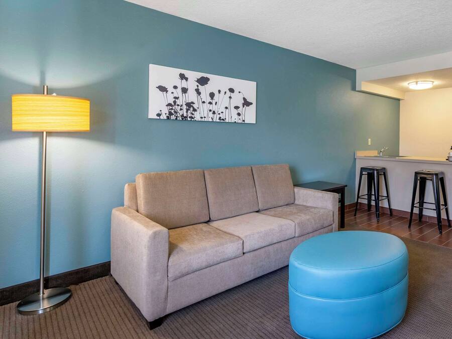 A living room with a beige sofa, a blue ottoman, a floor lamp, and a kitchen area with stools.