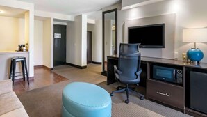 A hotel room with a desk, chair, television, microwave, and a small kitchenette.