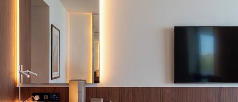Et moderne hotelværelse med en trækommode, et stort spejl, et fladskærms-TV på væggen og et natbord med en lampe.