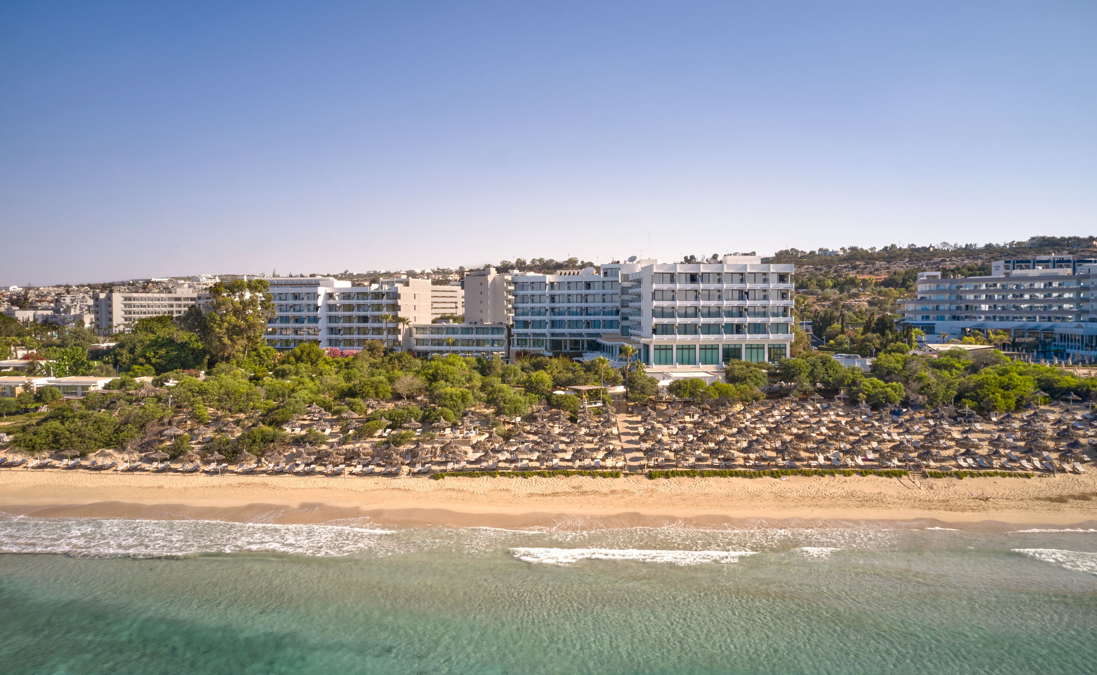 Un hotel frente al mar con varios pisos, una playa en primer plano y un paisaje urbano al fondo.