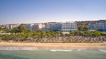 Un hotel frente al mar con varios pisos, una playa en primer plano y un paisaje urbano al fondo.
