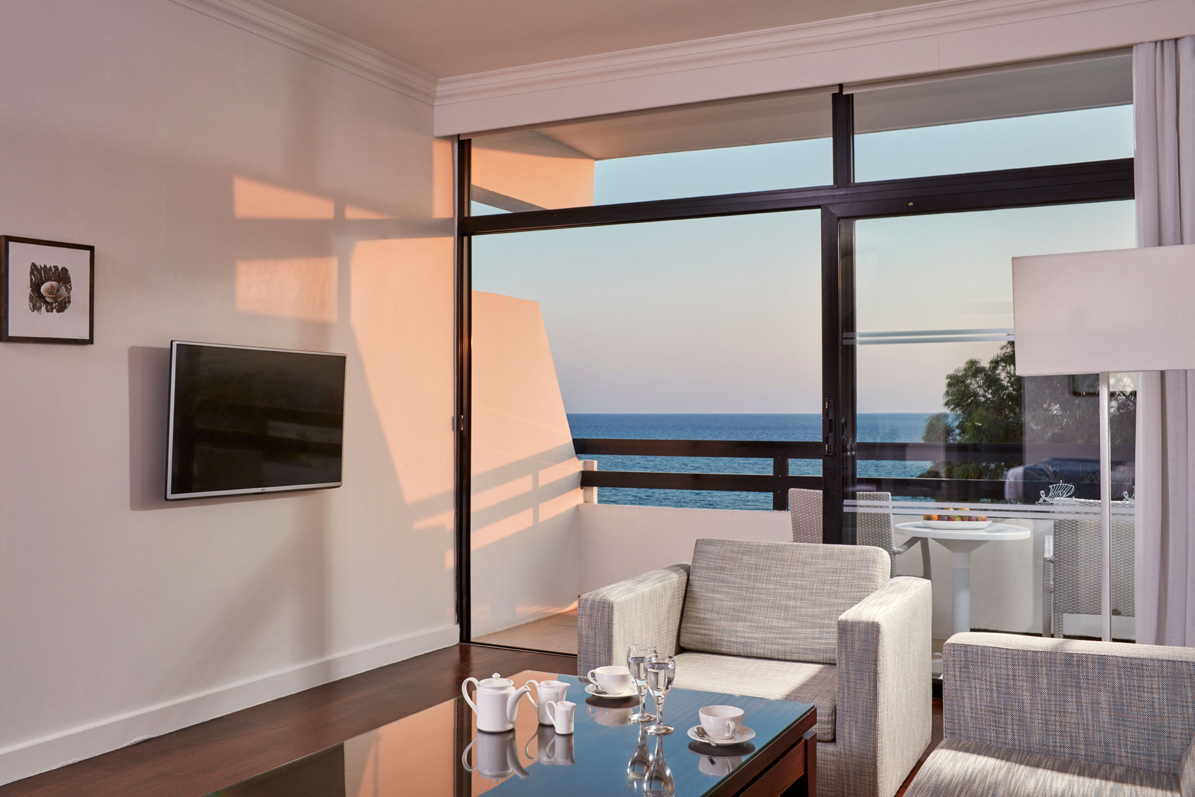 A modern hotel room with a balcony, a flat-screen TV, a dining table set with tea and glasses, and a view of the sea.