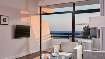A modern hotel room with a balcony, a flat-screen TV, a dining table set with tea and glasses, and a view of the sea.