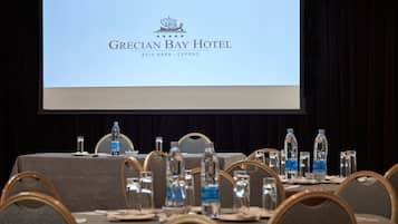 Une table de conférence, ornée de bouteilles d’eau, de verres et de plaques nominatives, est placée devant un écran de projection sur lequel est affiché le logo de l’hôtel Grecian Bay.