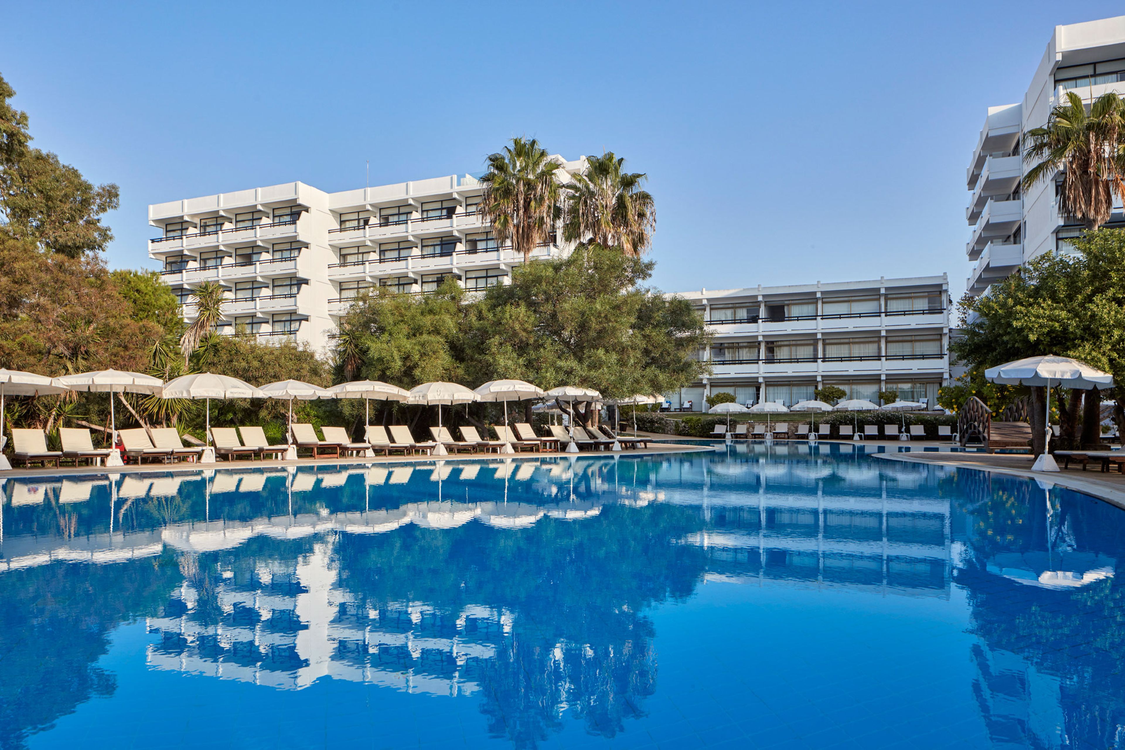 A swimming pool with lounge chairs and umbrellas, surrounded by multi-story buildings with balconies and palm trees.