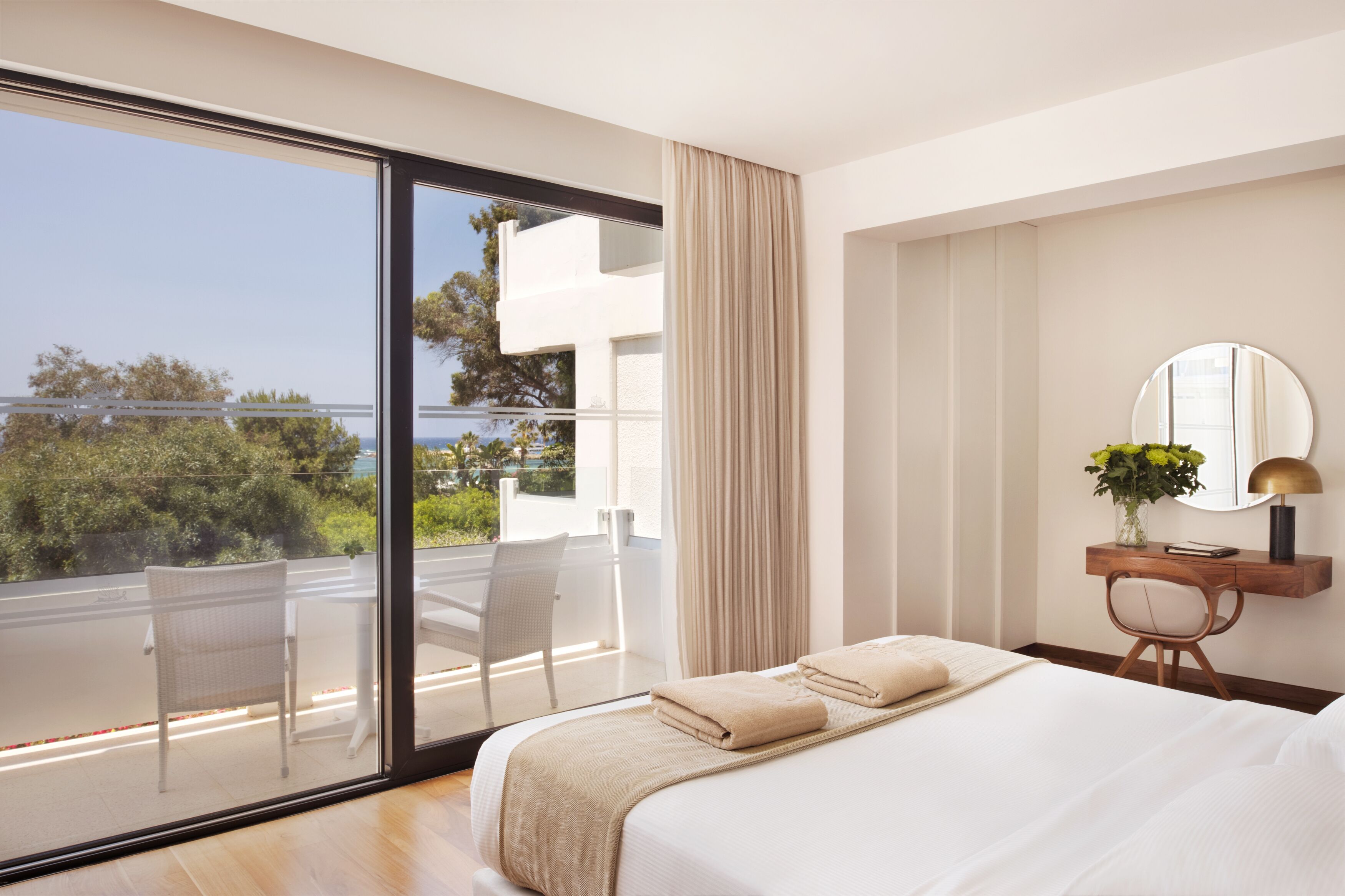 Una camera d'albergo moderna con un ampio balcone, un letto con asciugamani ordinatamente piegati, una scrivania in legno con uno specchio e vista sul mare.