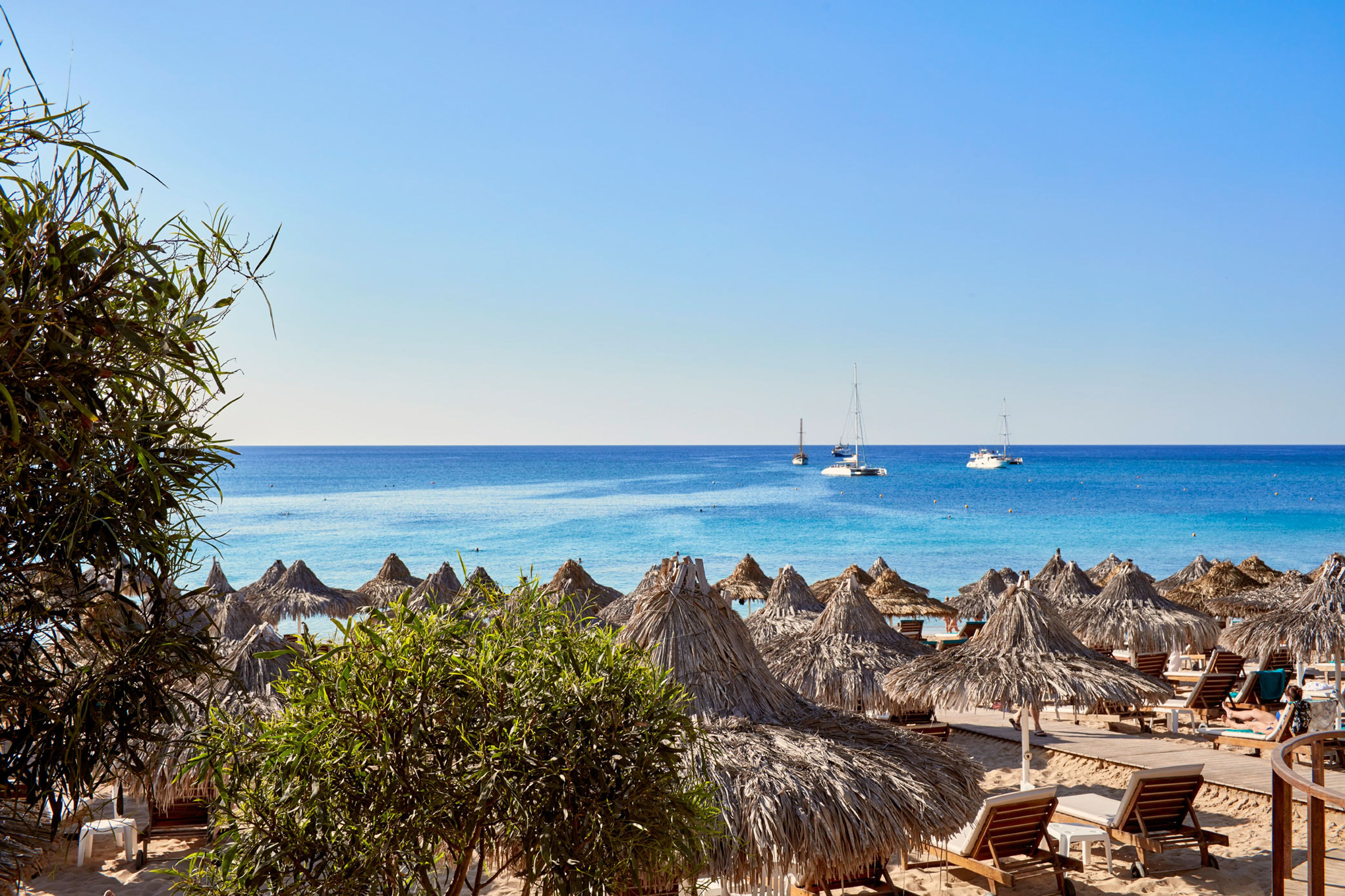 A beach with thatched umbrellas and wooden lounge chairs, a clear blue sea, and sailboats in the distance.