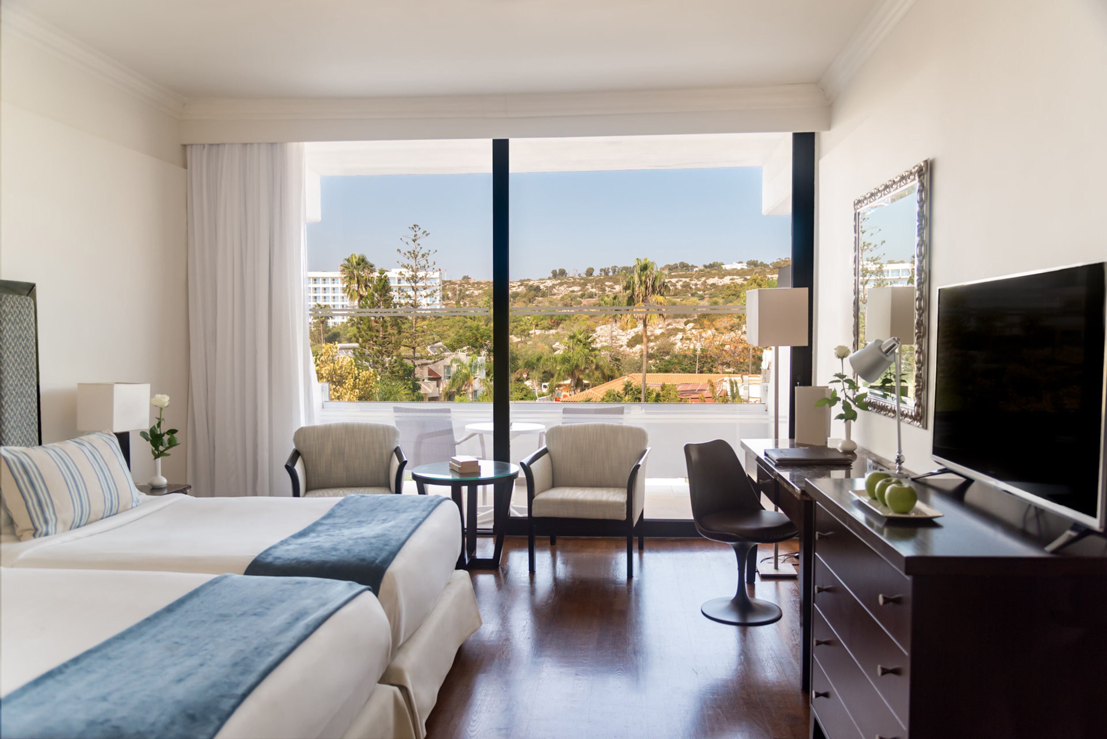 A hotel room with a large bed, two armchairs, a small table, a TV, and a view of a cityscape through large windows.