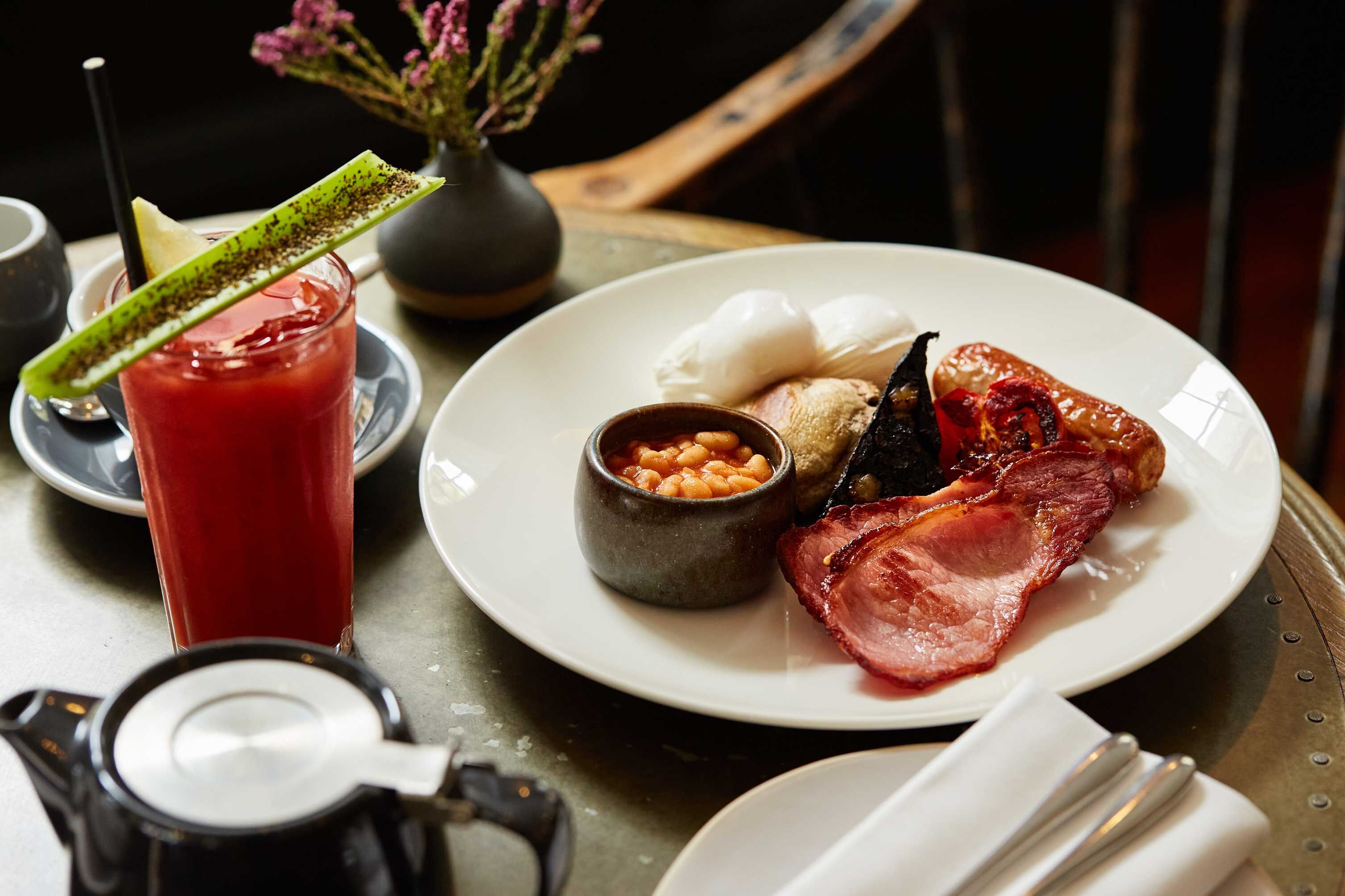 A plate with ham, beans, and a poached egg on a table with a coffee pot and a drink.