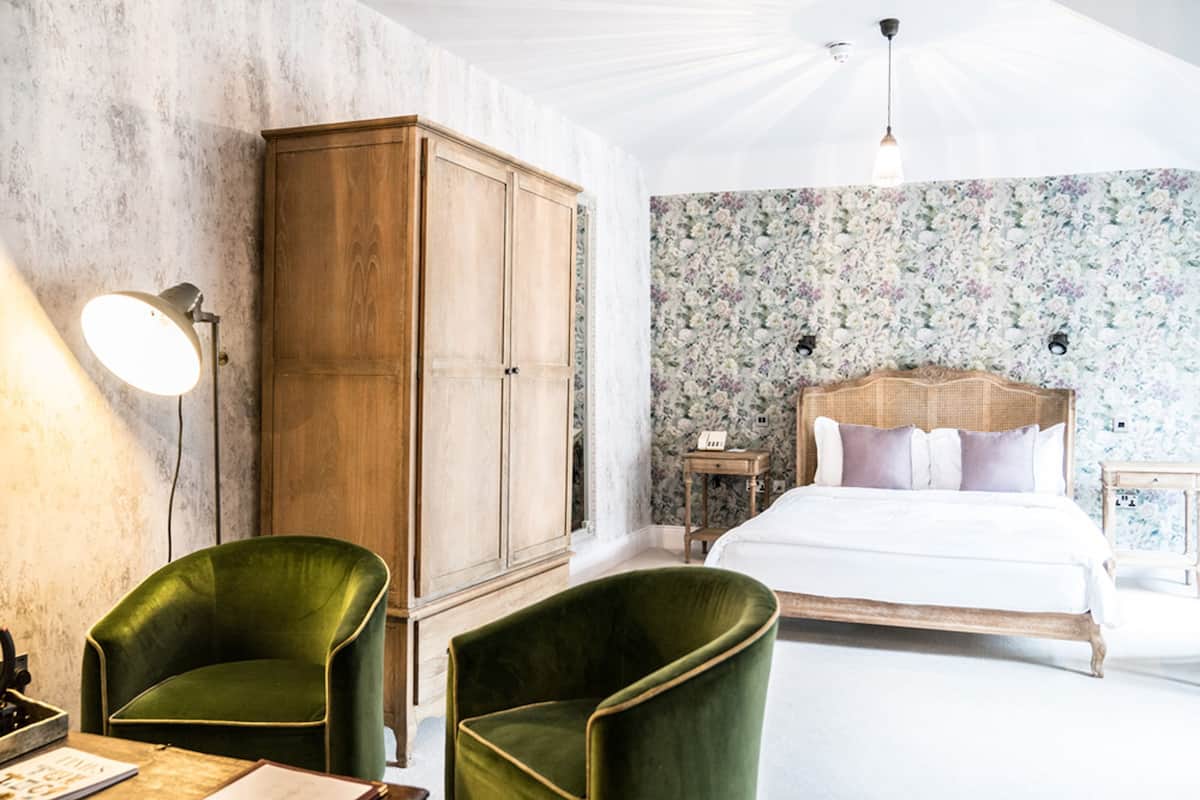 Quarto com um guarda-roupa de madeira grande, uma poltrona verde, uma cama com roupa de cama branca e papel de parede floral.