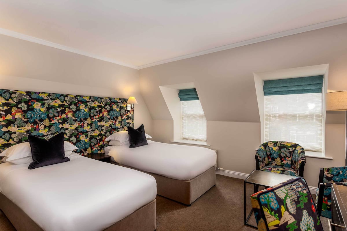 Quarto de hotel com duas camas, uma cadeira e uma mesinha.