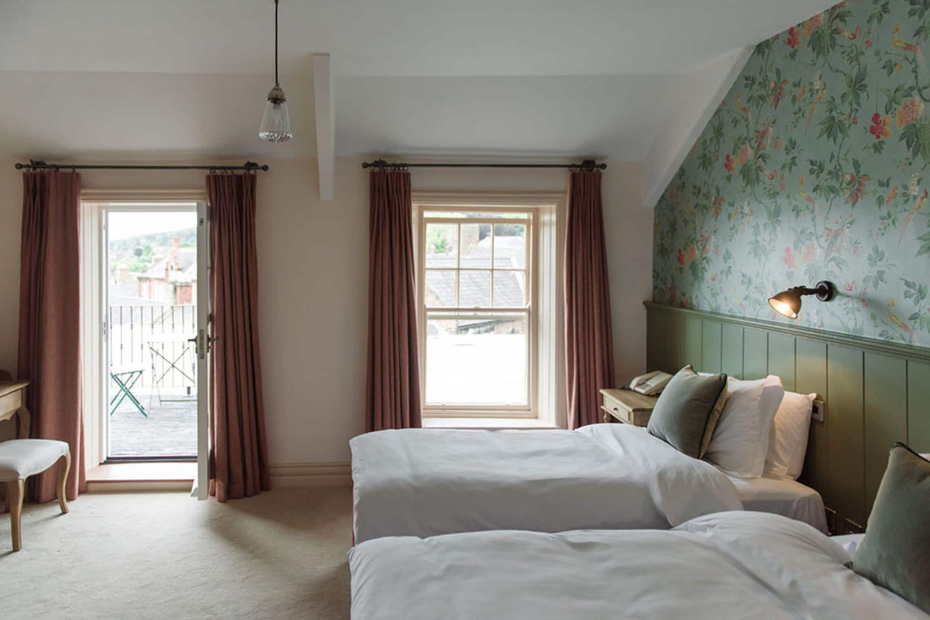 Een slaapkamer met twee bedden, een bureau en een stoel. Er zijn ramen met gordijnen, een wand met bloemenbehang en een hanglamp.