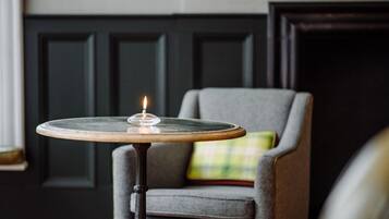 A round table with a candle, a grey armchair with a plaid cushion, and a dark wooden door in the background.