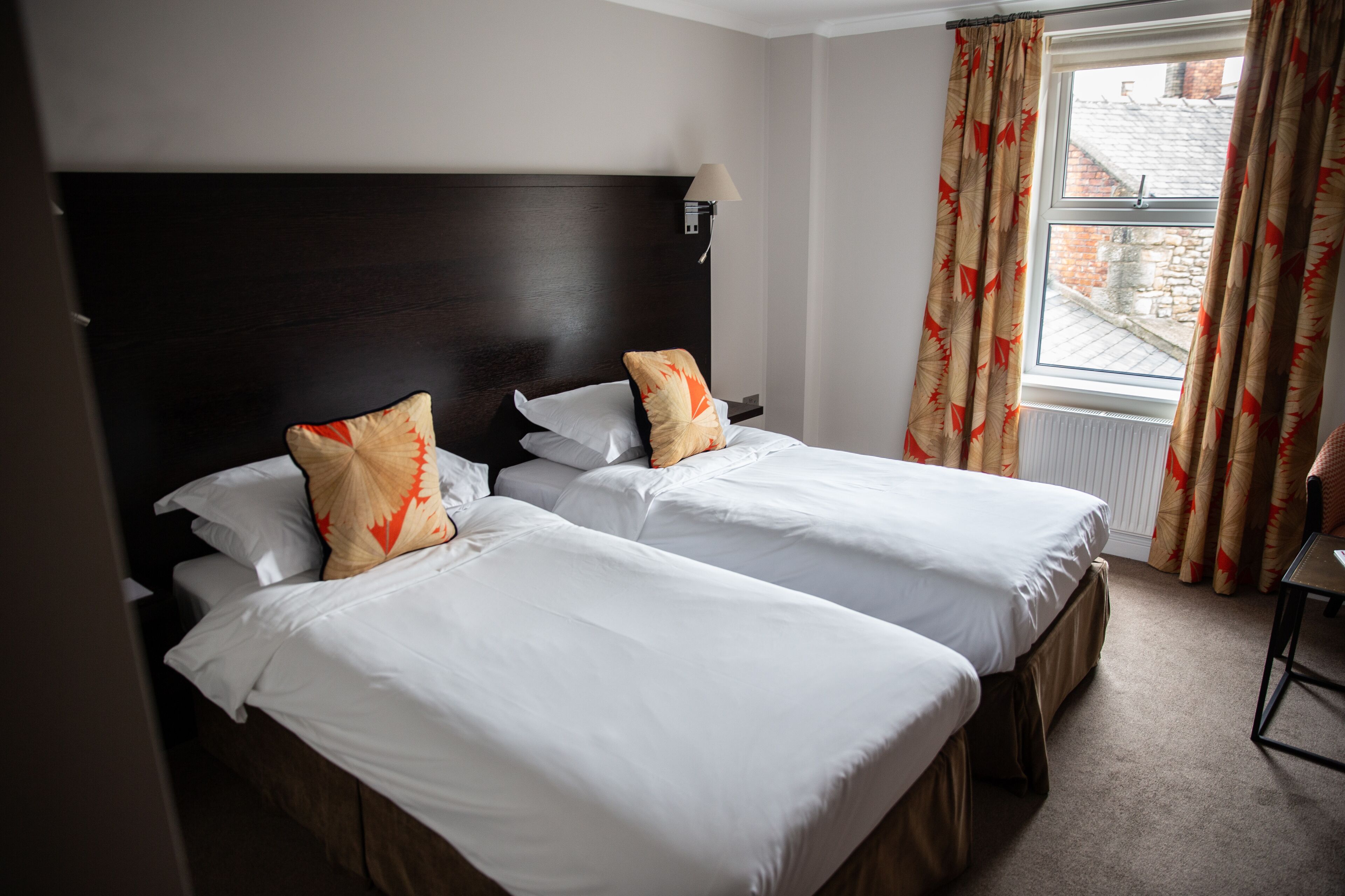 Une chambre d’hôtel avec deux lits simples, une tête de lit sombre, une fenêtre avec des rideaux et une petite table avec une chaise.