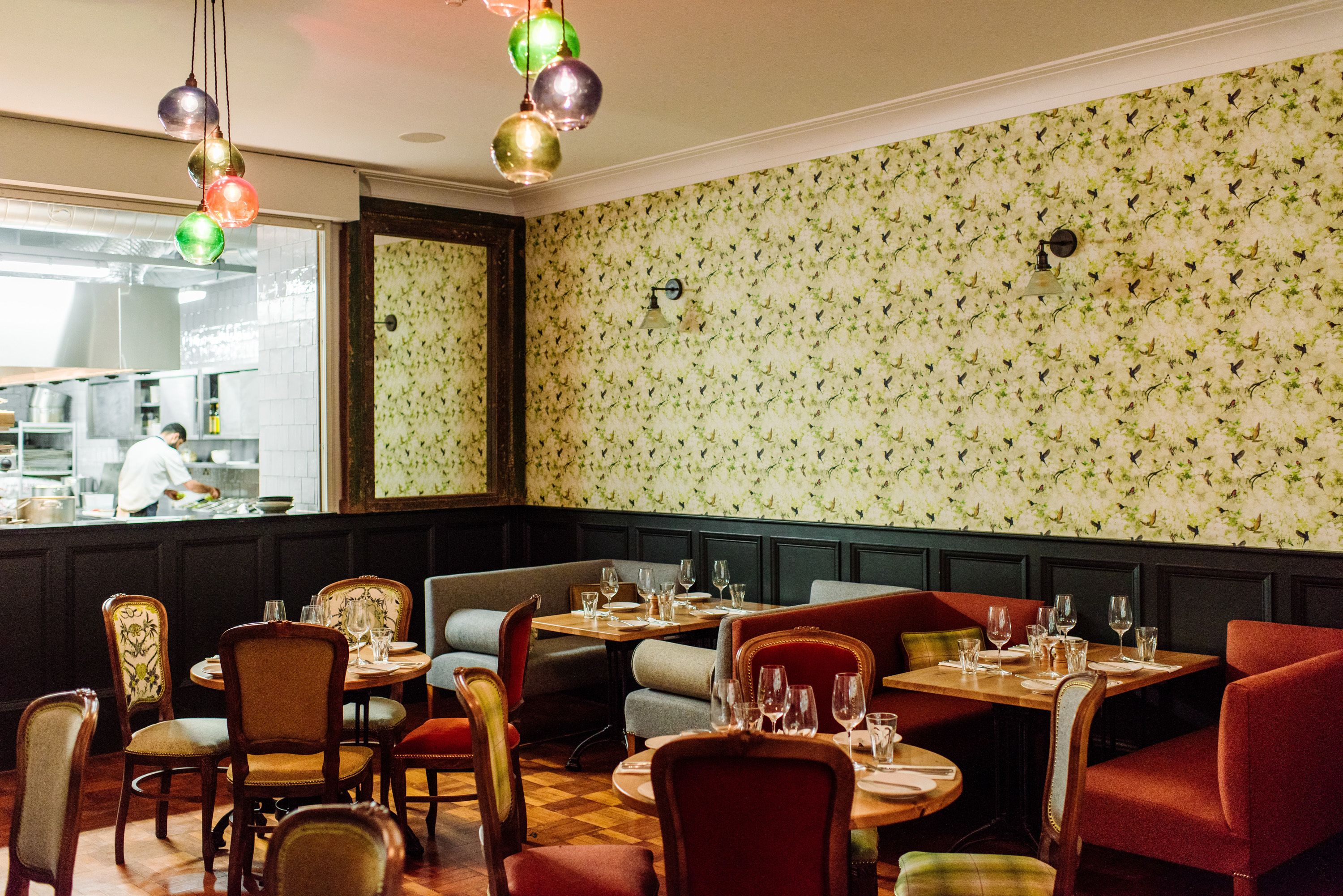 A restaurant with booths and tables, a kitchen in the background, and patterned wallpaper.