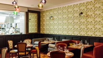 A restaurant with booths and tables, a kitchen in the background, and patterned wallpaper.