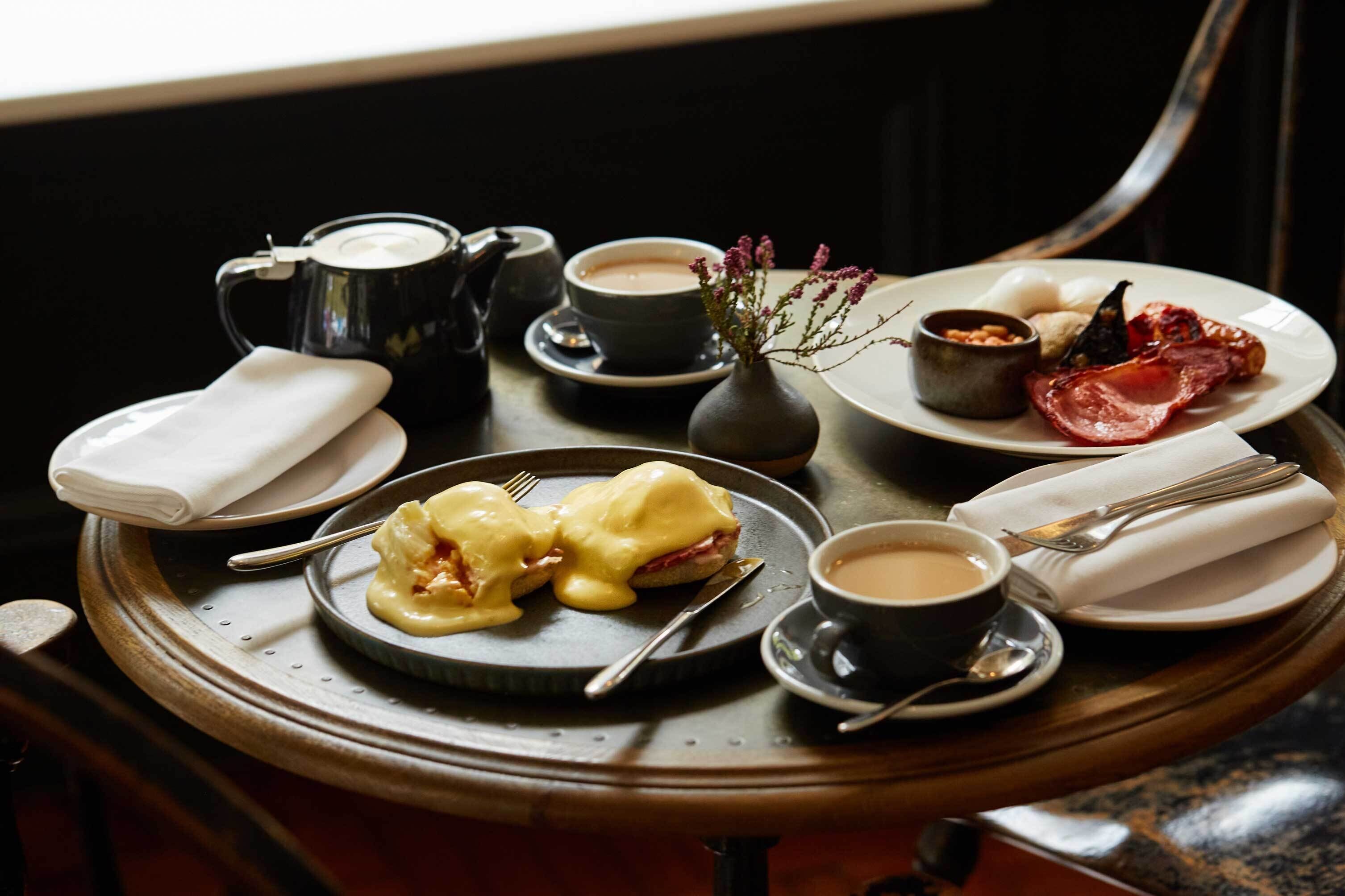Une table en bois dressée pour le petit-déjeuner, avec des œufs Bénédicte, du bacon, du café et du thé.