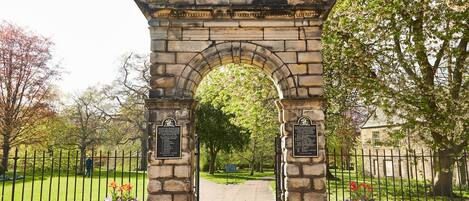 A stone archway with plaques, leading to a pathway surrounded by a fence and trees.