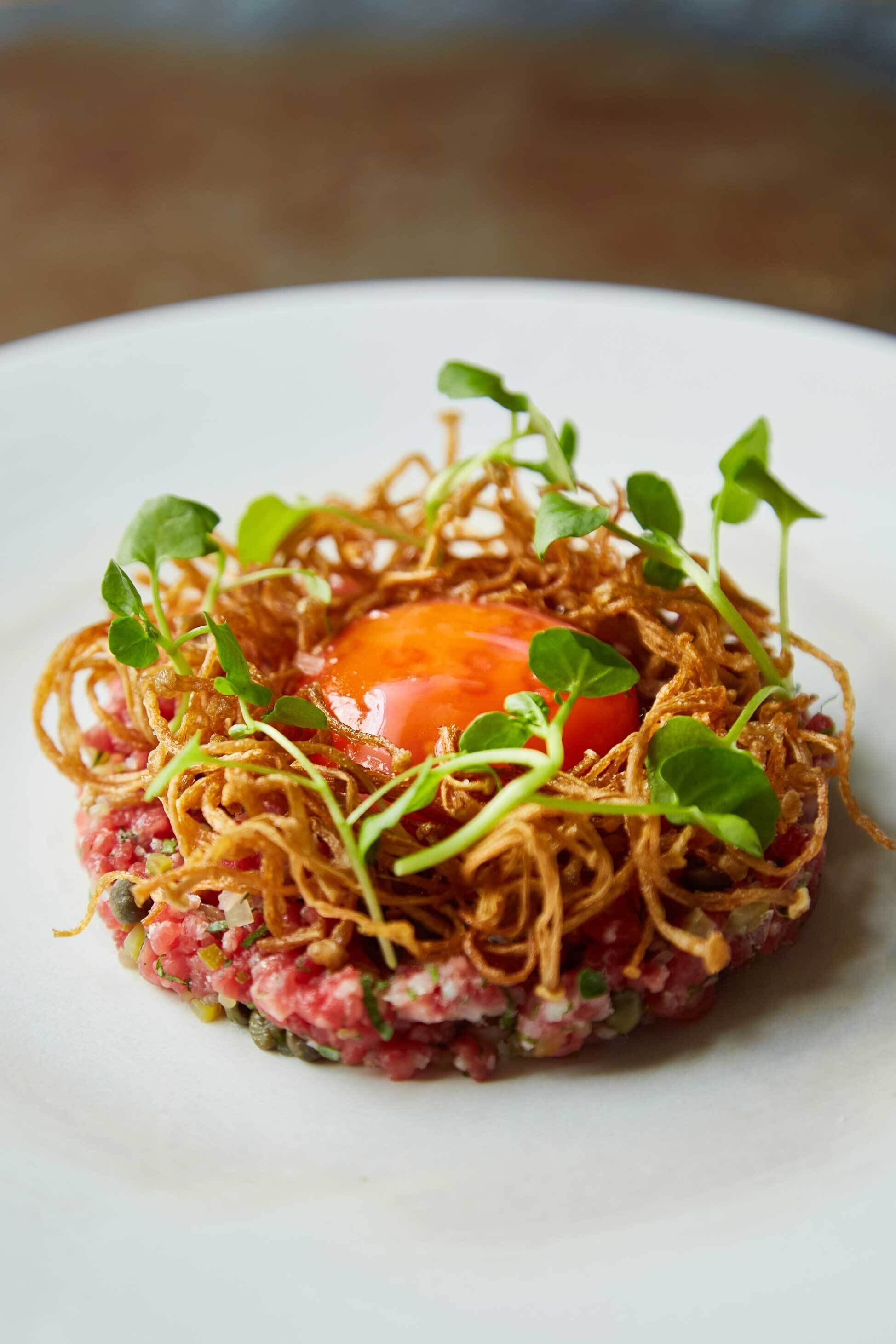 A dish with a raw meat center, garnished with microgreens and fried elements, on a white plate.