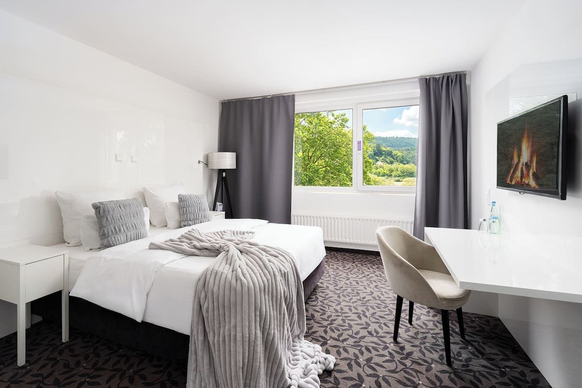 A hotel room with a bed, a desk, a chair, a lamp, a window with a view of greenery, and a fireplace.