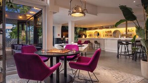 Ein modernes Interieur mit einer Essecke mit runden Tischen und farbenfrohen Stühlen, einer Bar mit einem runden Spiegel und Blick durch große Fenster.
